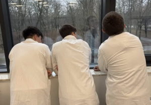 Uczniowie w fartuchach stoją tyłem przy oknie